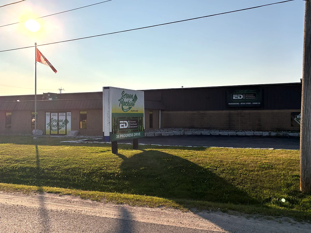 35 Progress Drive from the road. A tall sign by the kerb reads Screen Machine
      Promotions above a green panel carrying the EDI logo and the words 35 Progress Drive.
      Behind it is a long brown building with a Canadian flag, and a sign on the wall reading
      warehousing, retail store, ediinc.ca.